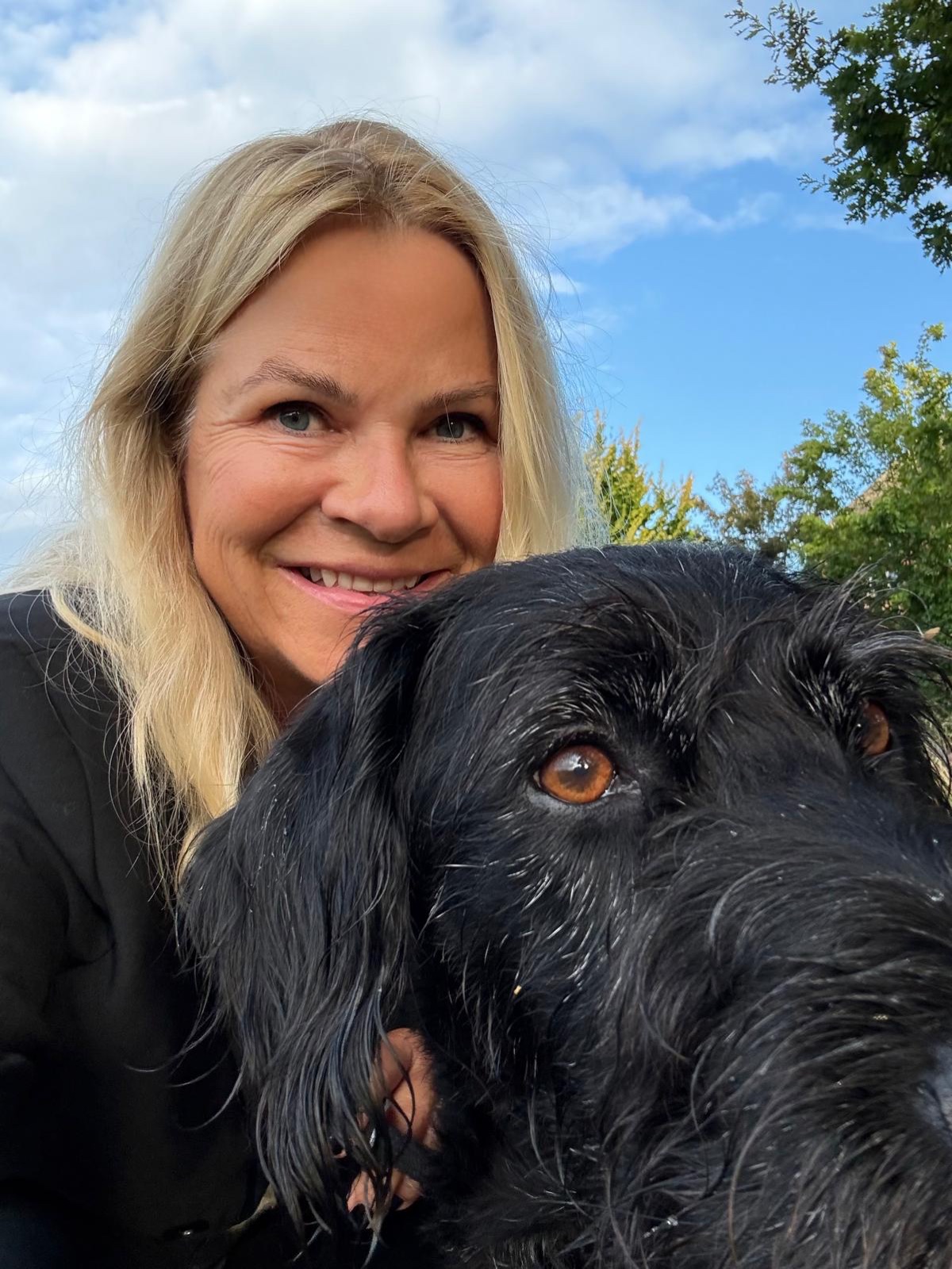 Ines Thordsen lächelnd mit ihrem schwarzen Hund unter blauem Himmel